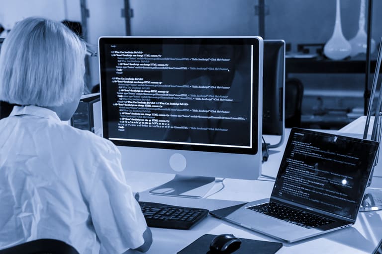 Pessoa de costas programando em uma estação de trabalho com dois monitores exibindo código